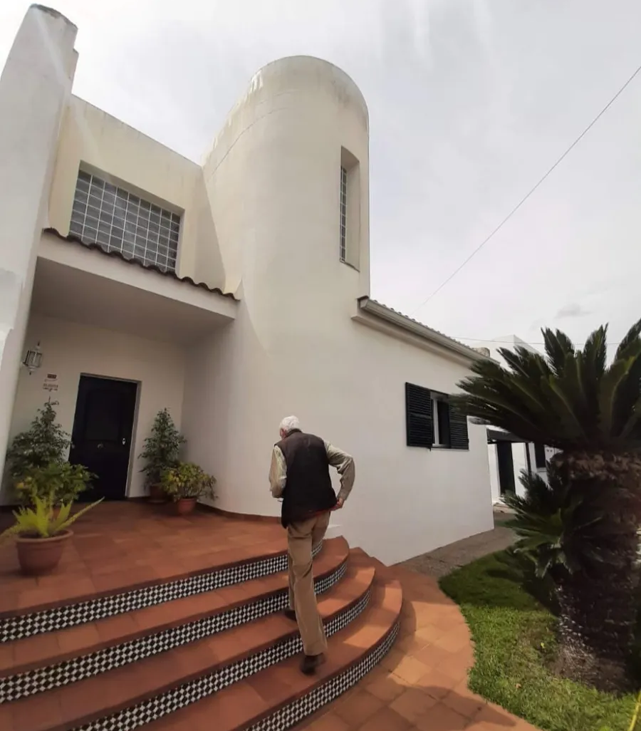 Una persona sube unos escalones de baldosas hacia una casa blanca con una torre redondeada y una palmera al frente.