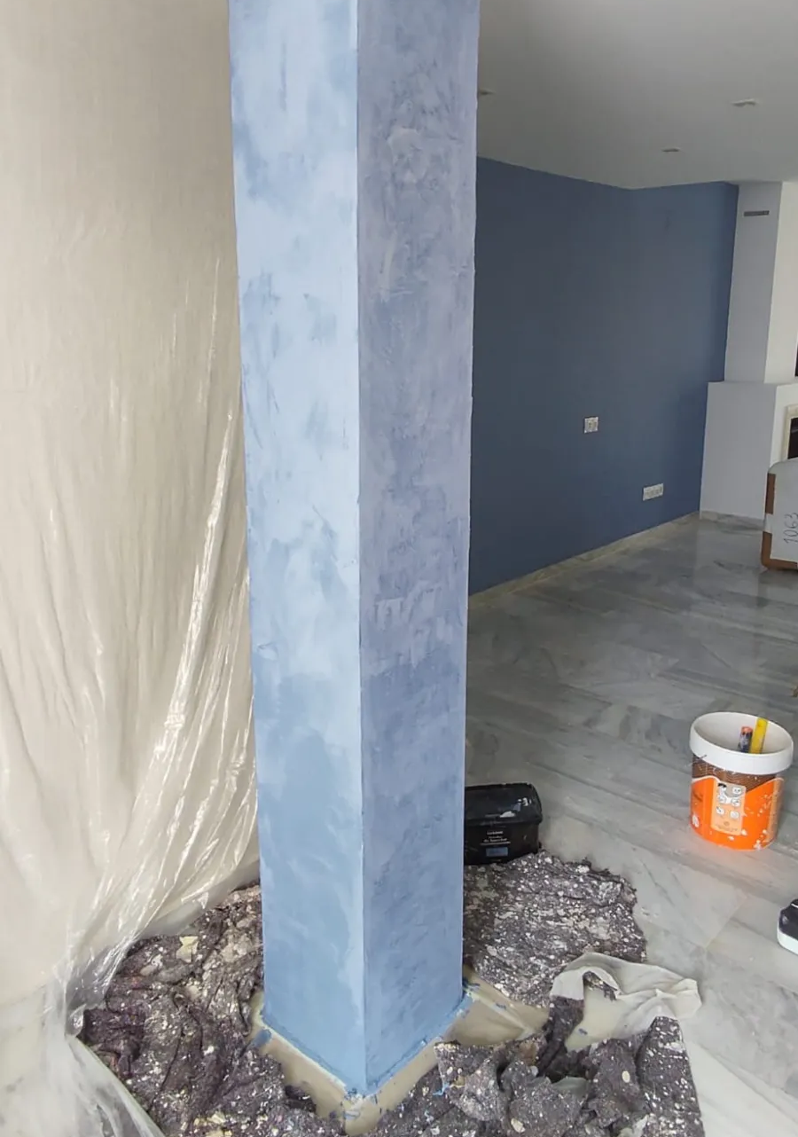 Habitación interior en obras de renovación con columna pintada de azul, pared cubierta de plástico, lona en el suelo y cubo de pintura.
