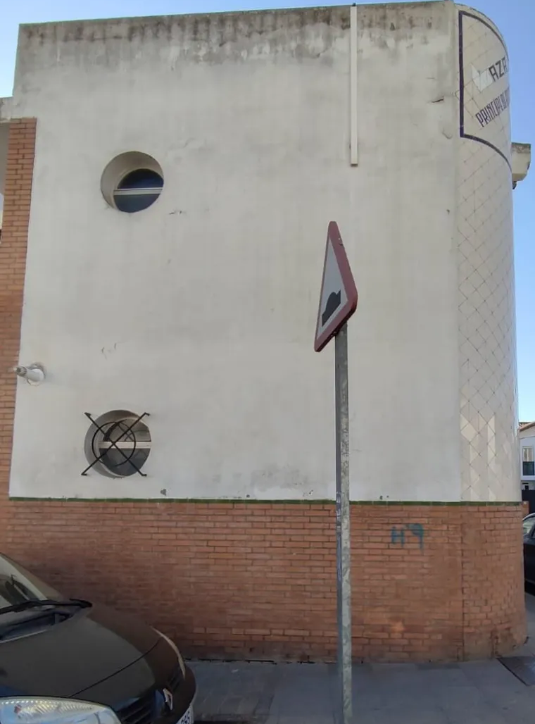Edificio de ladrillo blanco con ventanas redondas detrás de una señal de calle y un coche aparcado, con base de ladrillo rojo.