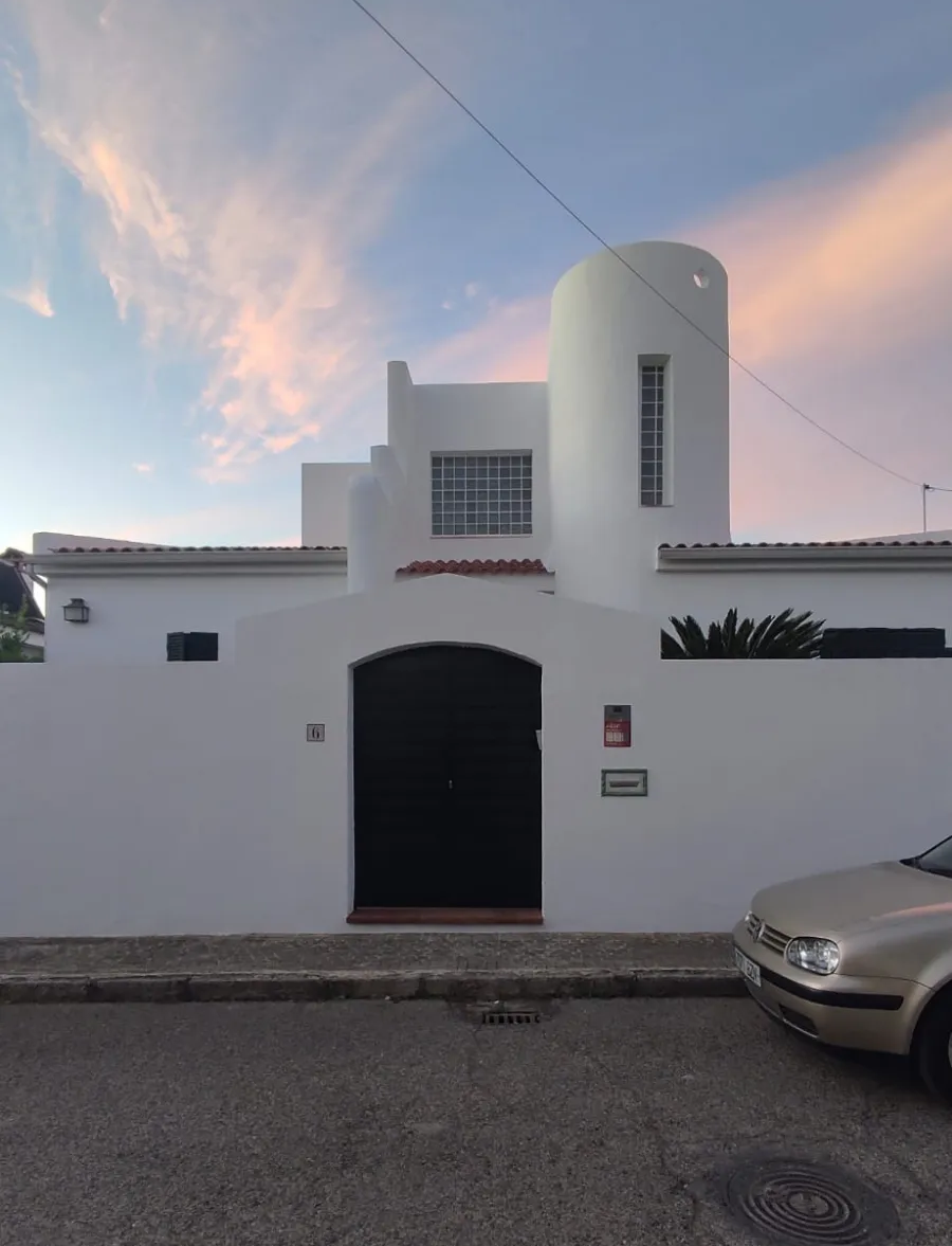 Una casa blanca con una verja negra al anochecer, nubes rosadas en el cielo y un coche plateado aparcado en la acera.