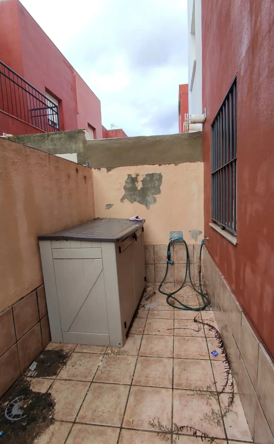 Estrecho nicho de servicio exterior con suelo de baldosas, un pequeño armario, manguera de pared y pintura descascarada en la pared del fondo.