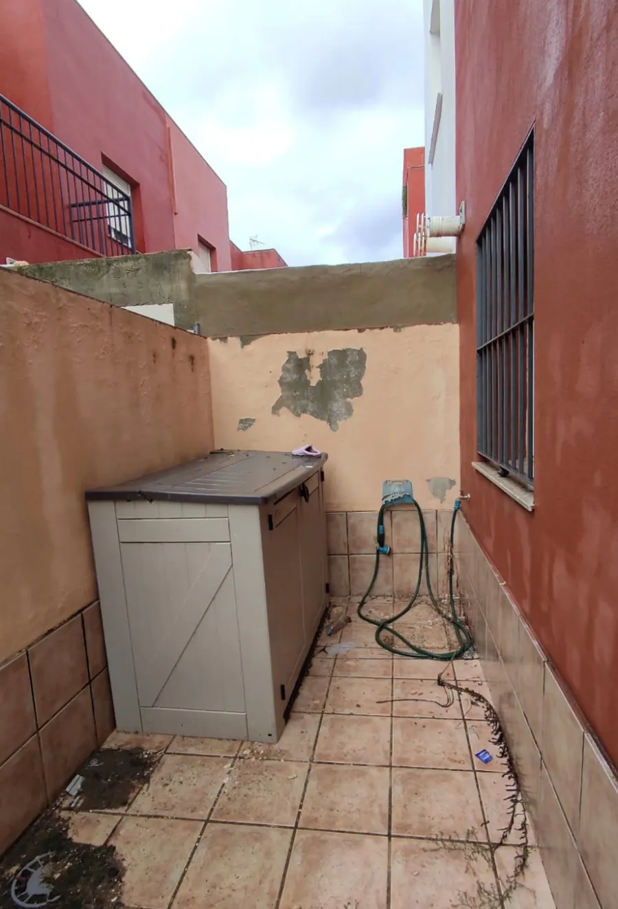 Patio exterior estrecho con suelo de baldosas, paredes de color beige, un armario de almacenamiento y una manguera a la derecha.