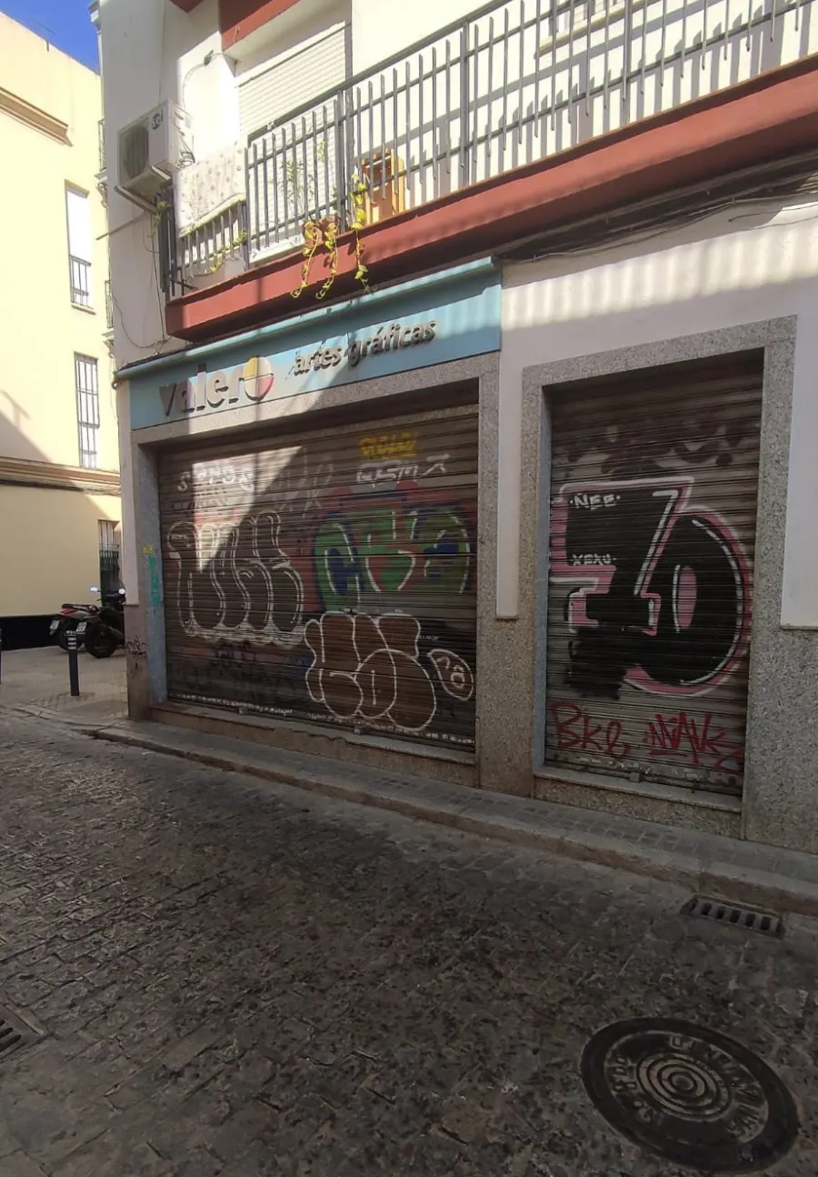 Escaparate cerrado con contraventanas metálicas cubiertas de grafitis en una estrecha calle empedrada, visto desde la distancia.
