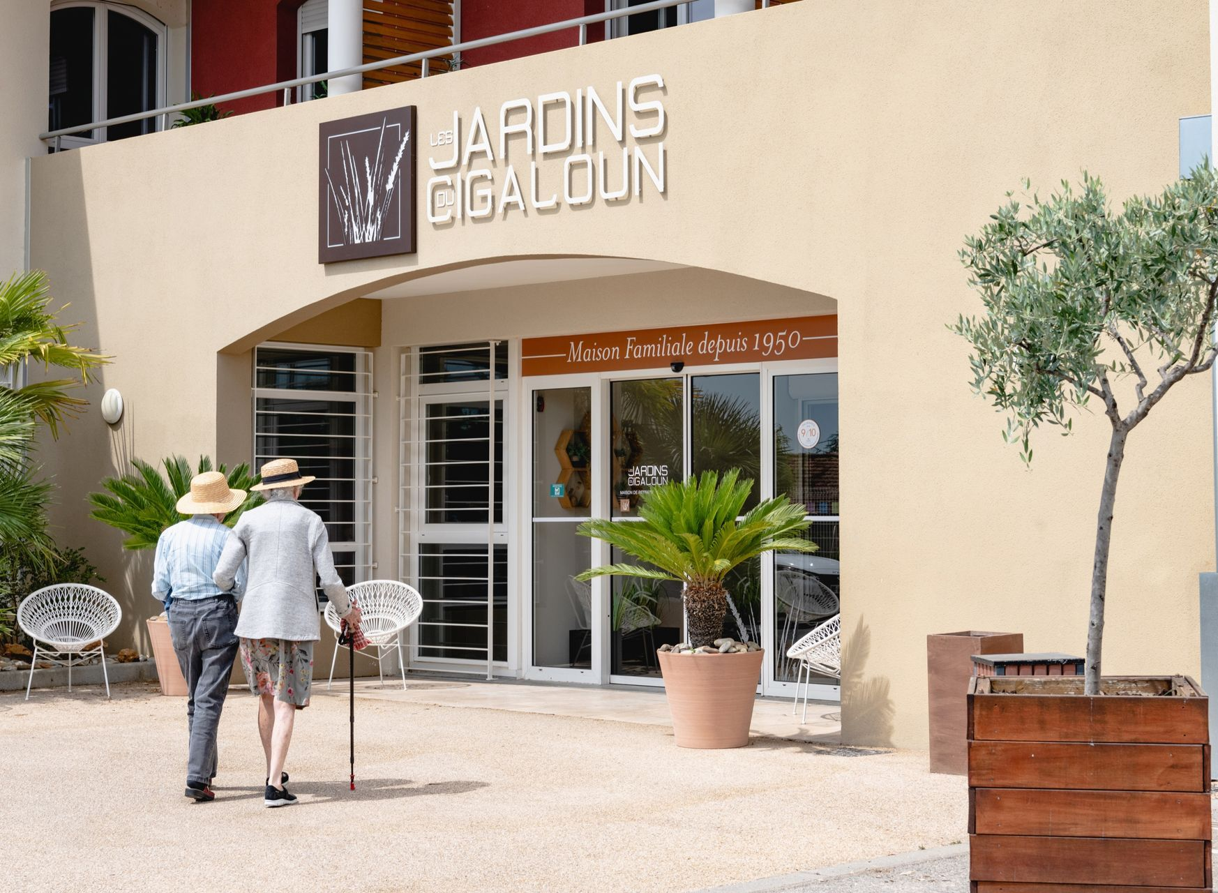 Deux personnes se dirigent vers l'entrée des Jardins du Cigaloun, un bâtiment beige orné de plantes en pot.
