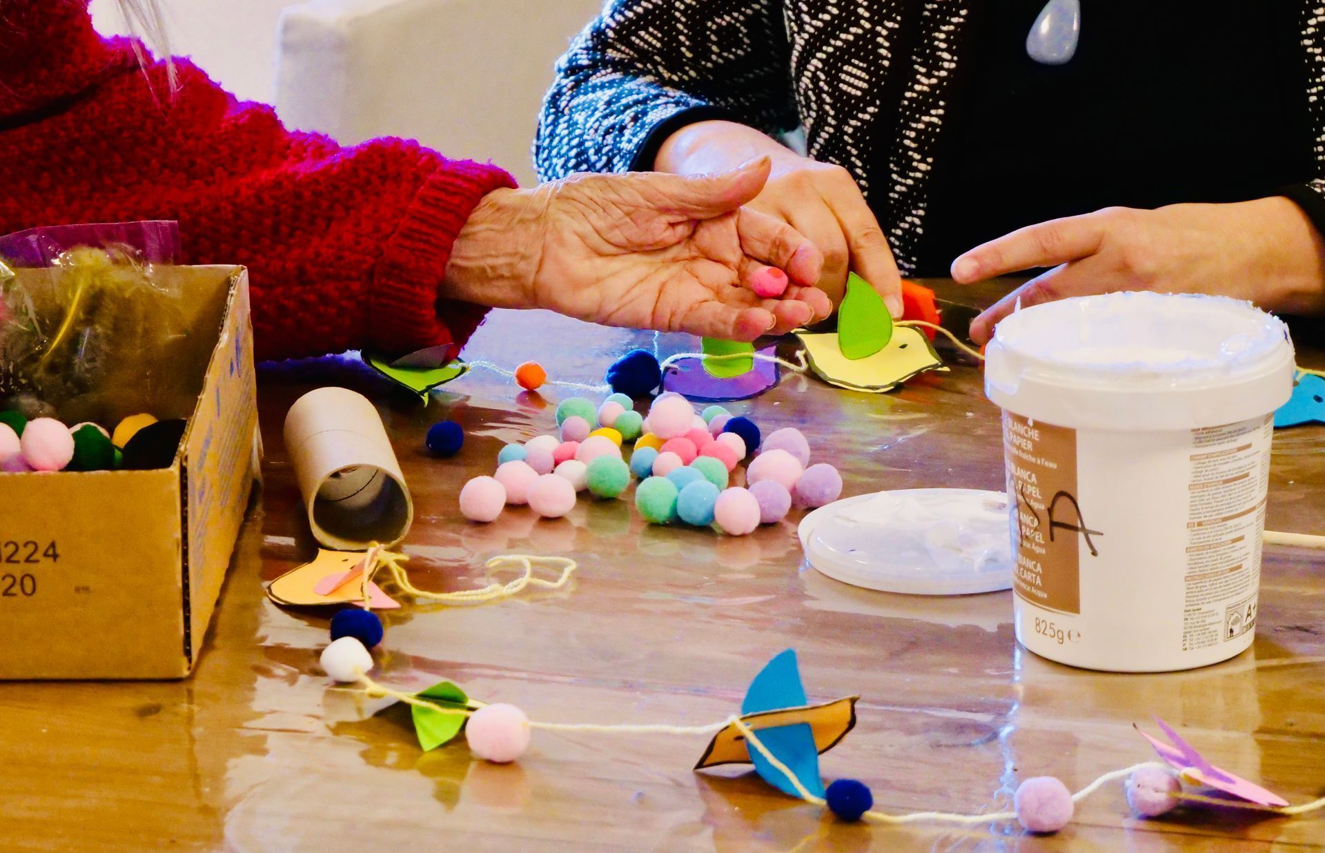 Des mains disposent des pompons colorés et des formes en papier sur une table avec un récipient blanc et une boîte en carton.