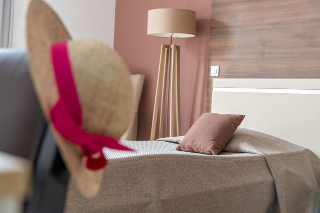 Chambre moderne avec murs roses, lit, lampadaire et un chapeau de paille flou au premier plan.