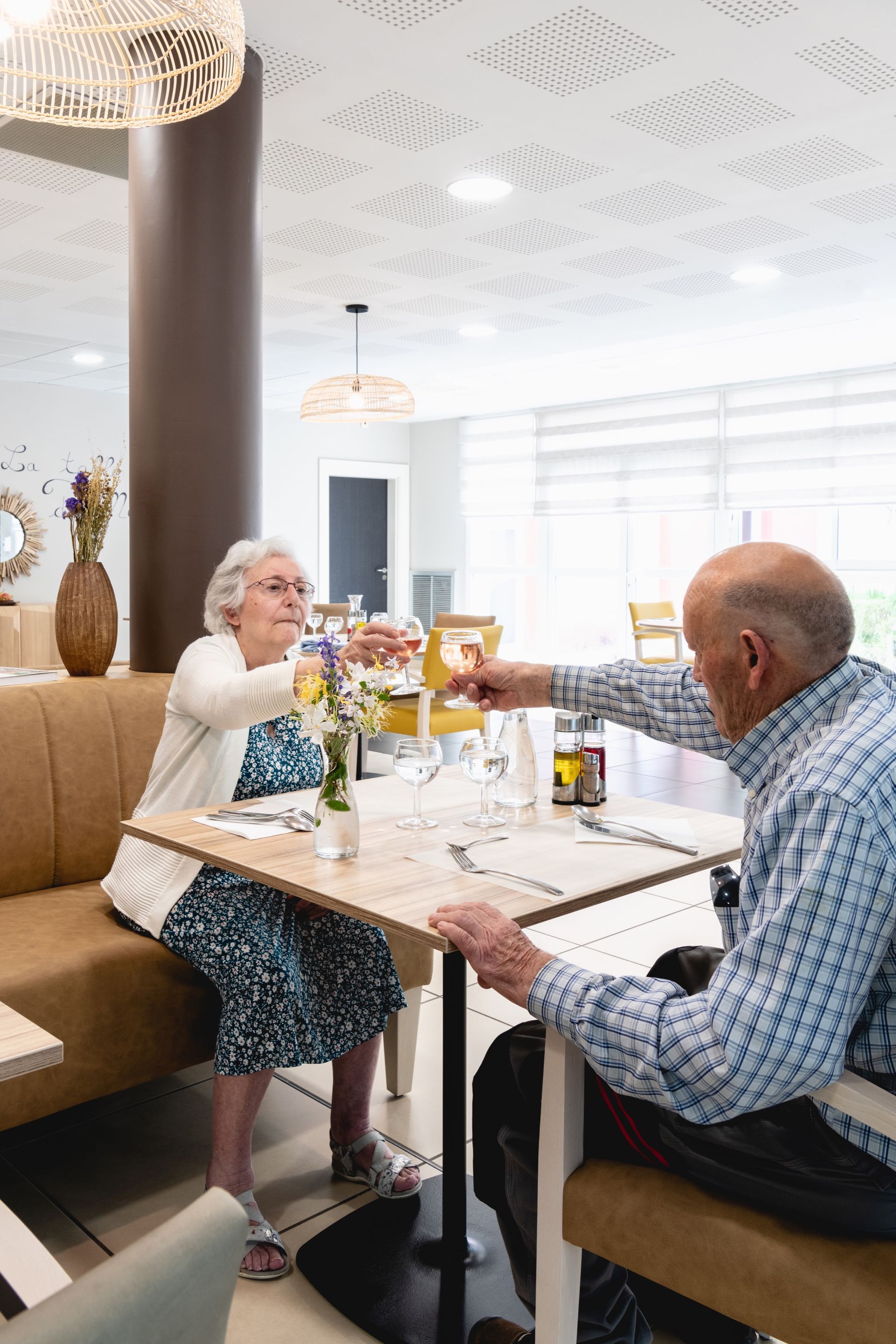Deux personnes trinquent à la terrasse d'un restaurant, assises dans un box lumineux avec des boissons et des fleurs.