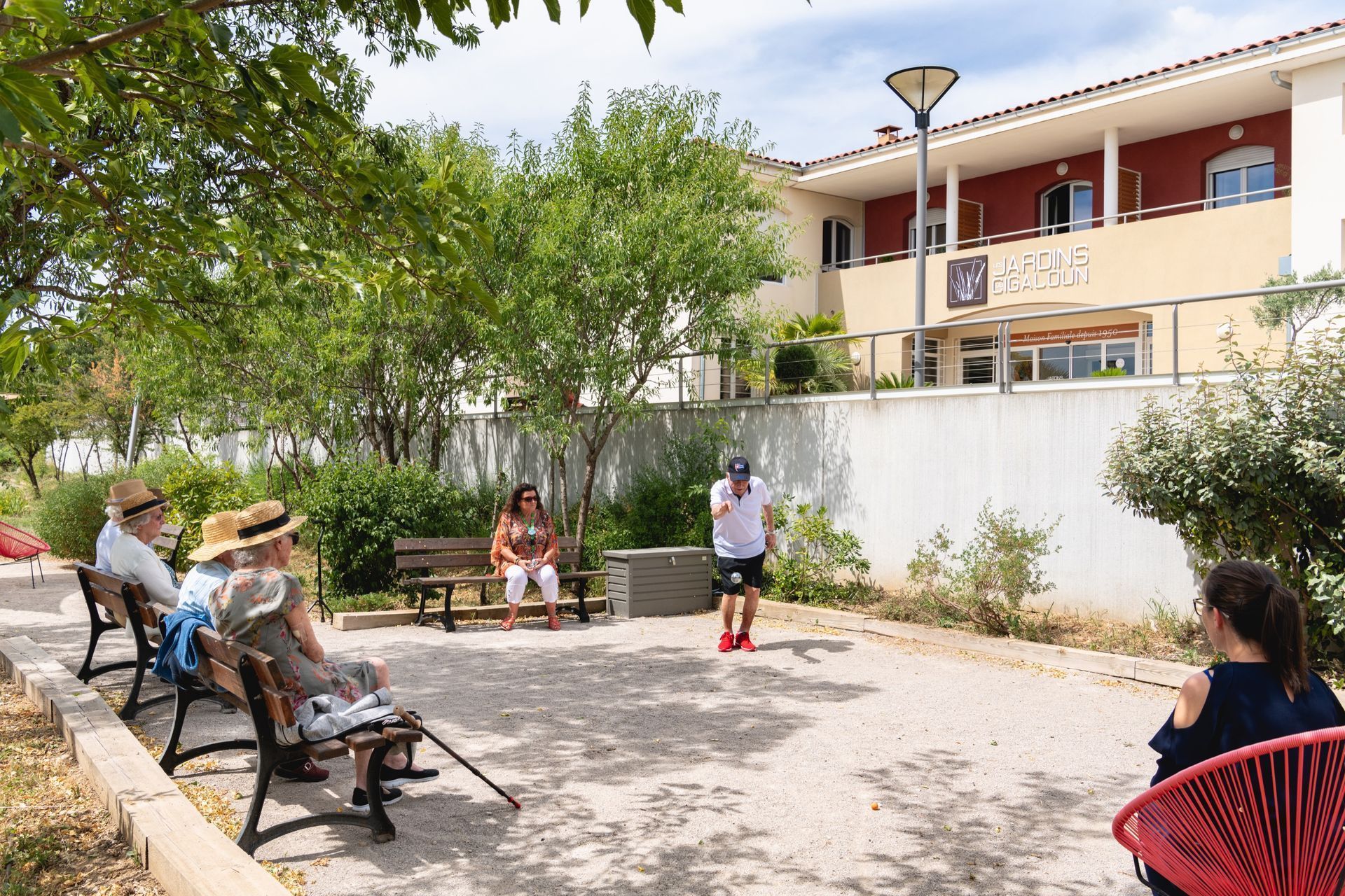 Des personnes assises autour d'une cour avec des bancs, des arbres et un bâtiment en arrière-plan.