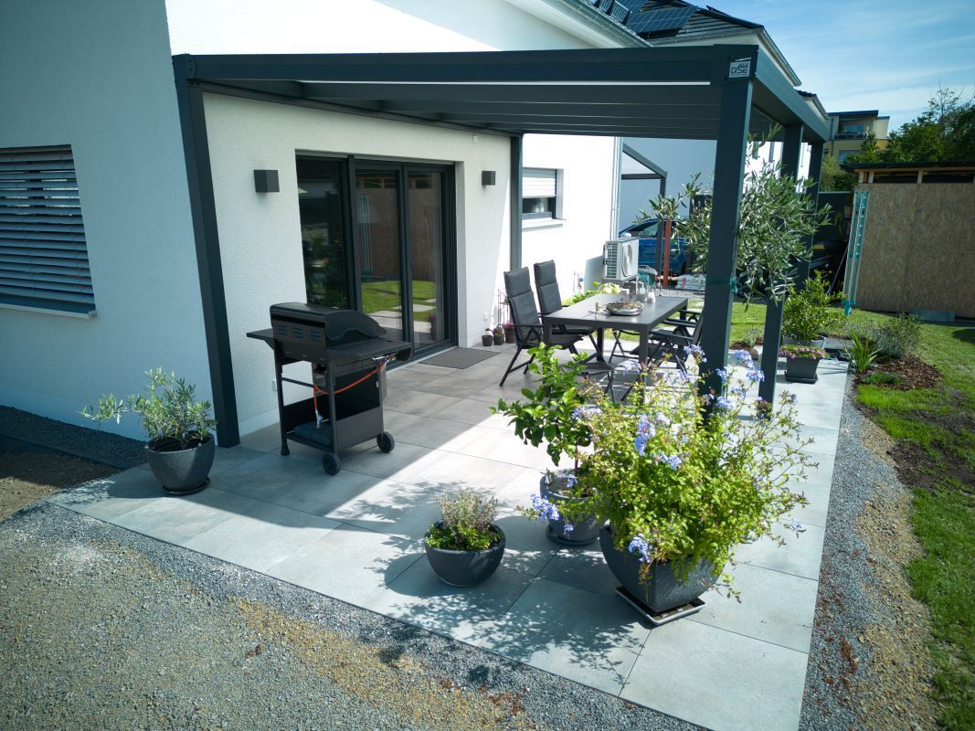 Eine Terrasse mit Tisch und Stühlen unter einer Pergola vor einem Haus.