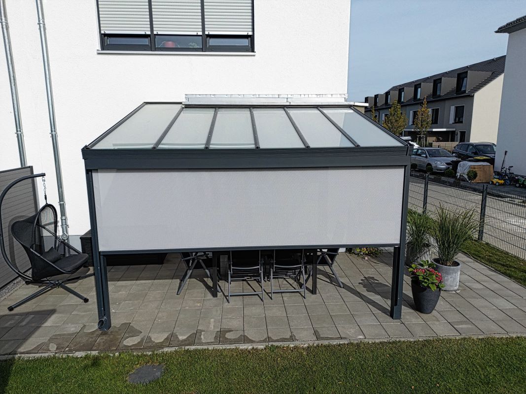 Eine Terrasse mit Tisch und Stühlen unter einem Vordach vor einem Haus.