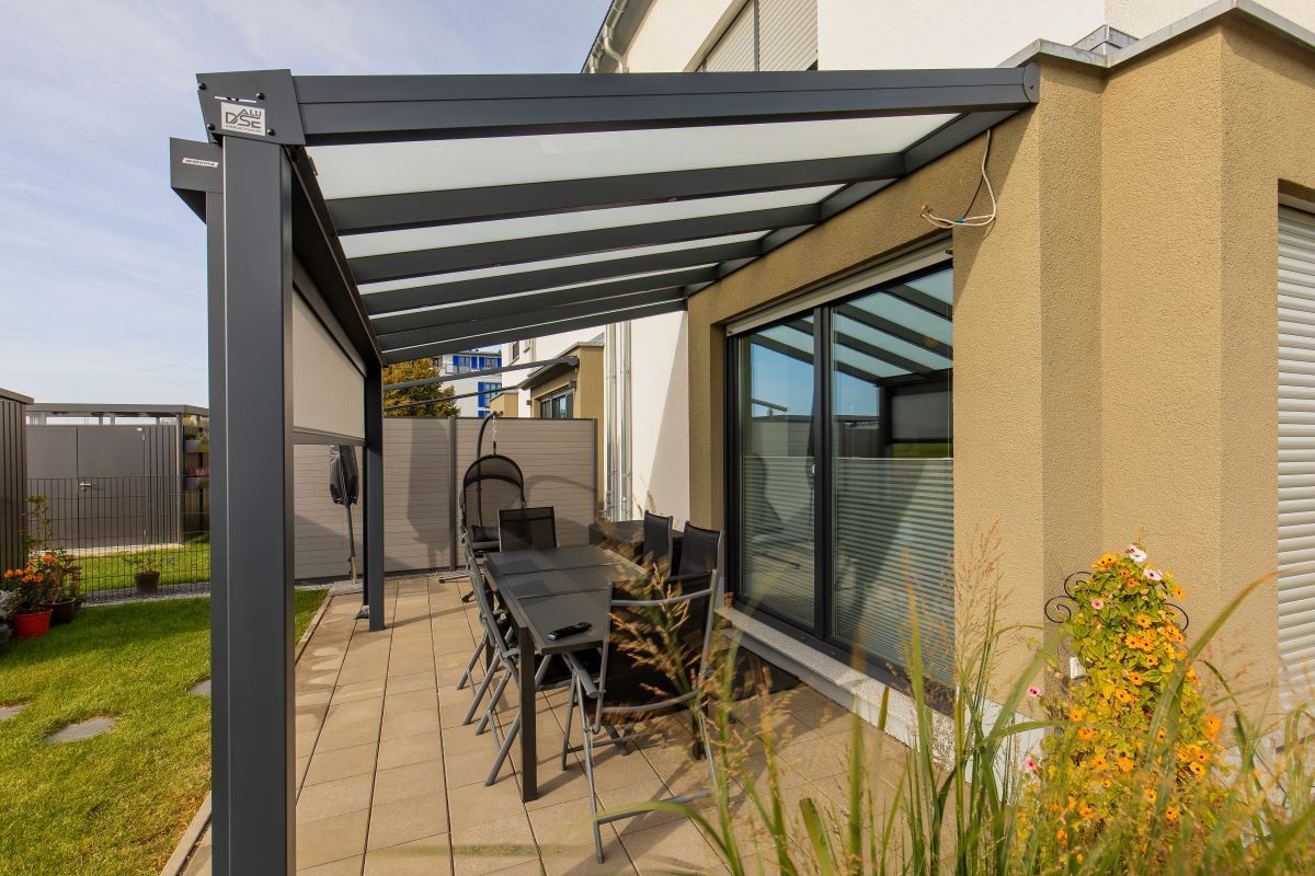 Eine Terrasse mit Tisch und Stühlen unter einem Vordach vor einem Haus.