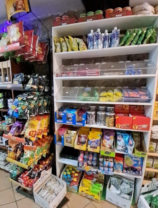 Pequeño escaparate de una tienda con coloridas patatas fritas, caramelos y artículos de aseo apilados en estantes.