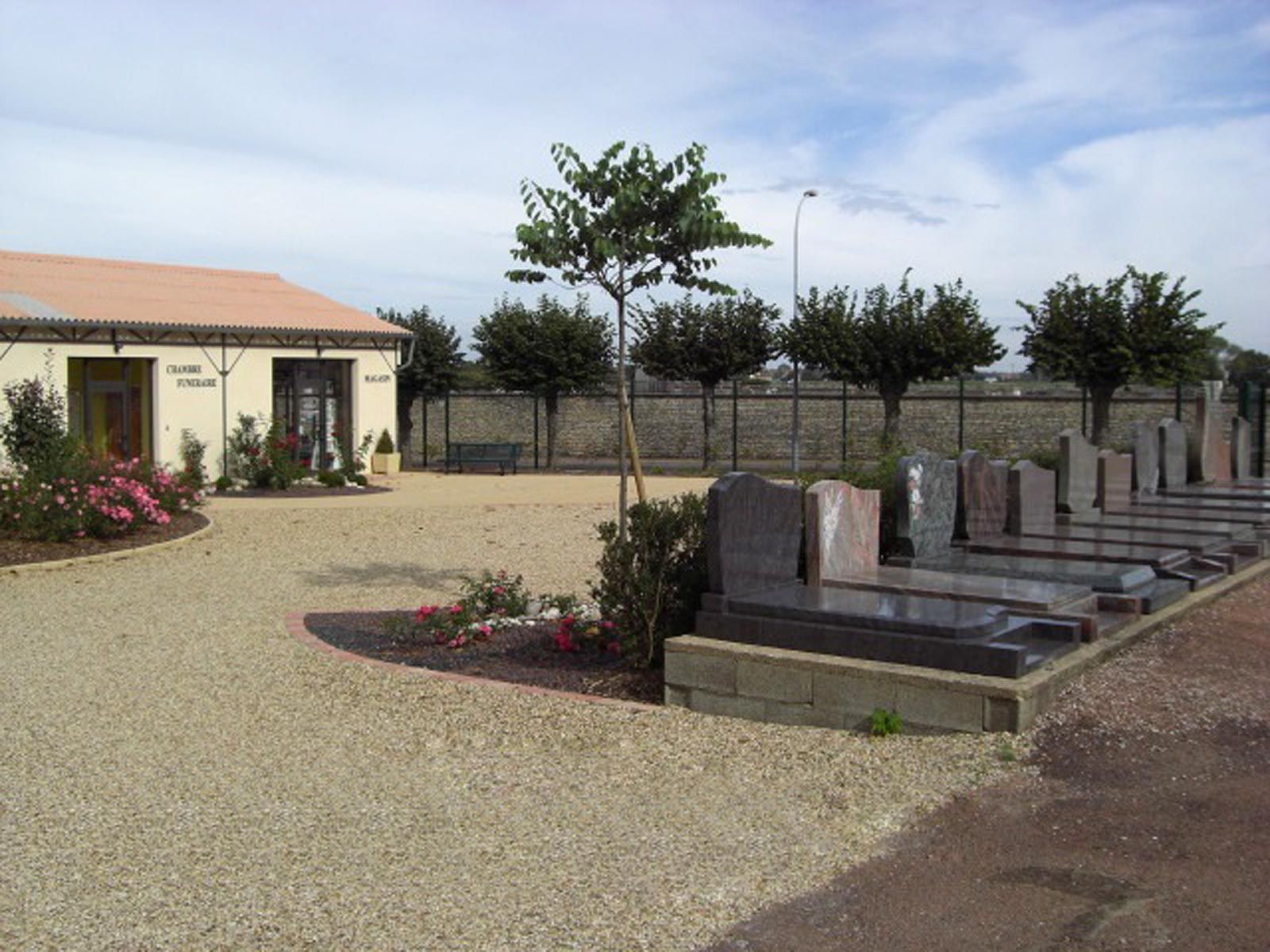 Mémorial extérieur avec plaques de pierre, un jeune arbre et un petit bâtiment sur un terrain gravillonné.