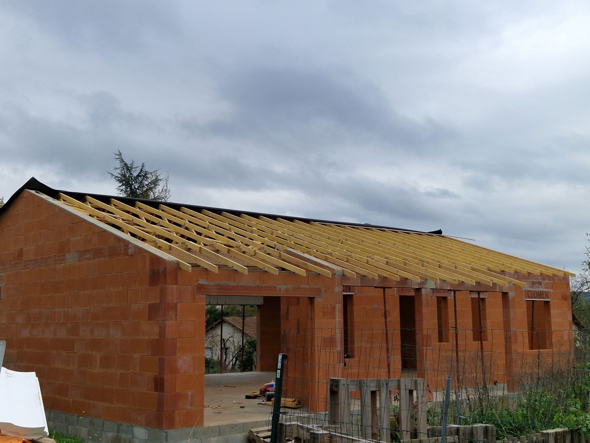 Une maison en briques orange partiellement construite, avec une charpente de toit en bois, sous un ciel nuageux et couvert.
