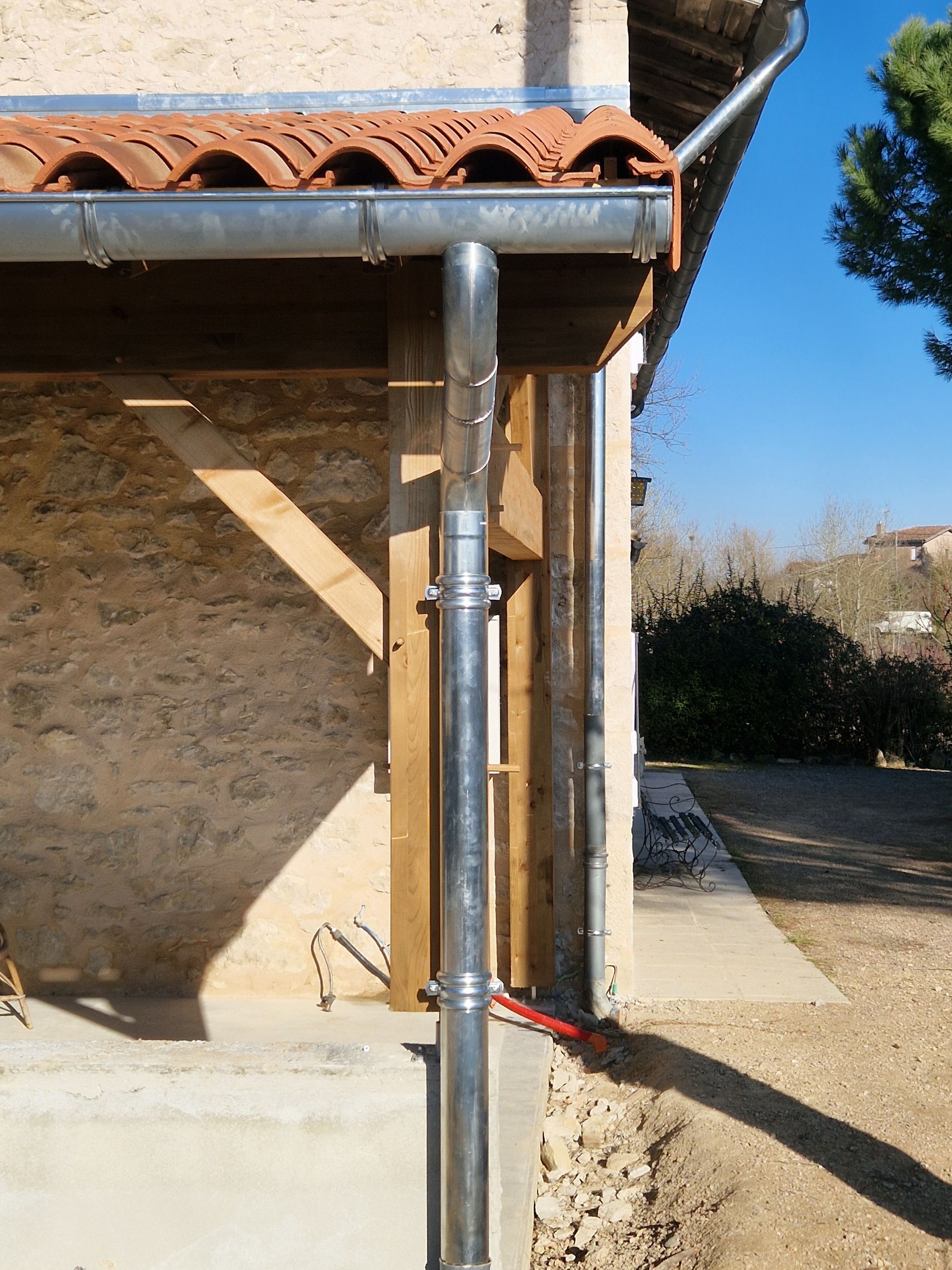 Une gouttière et une descente pluviale en métal fixées à l'angle d'un bâtiment en pierre avec un toit en tuiles de terre cuite.