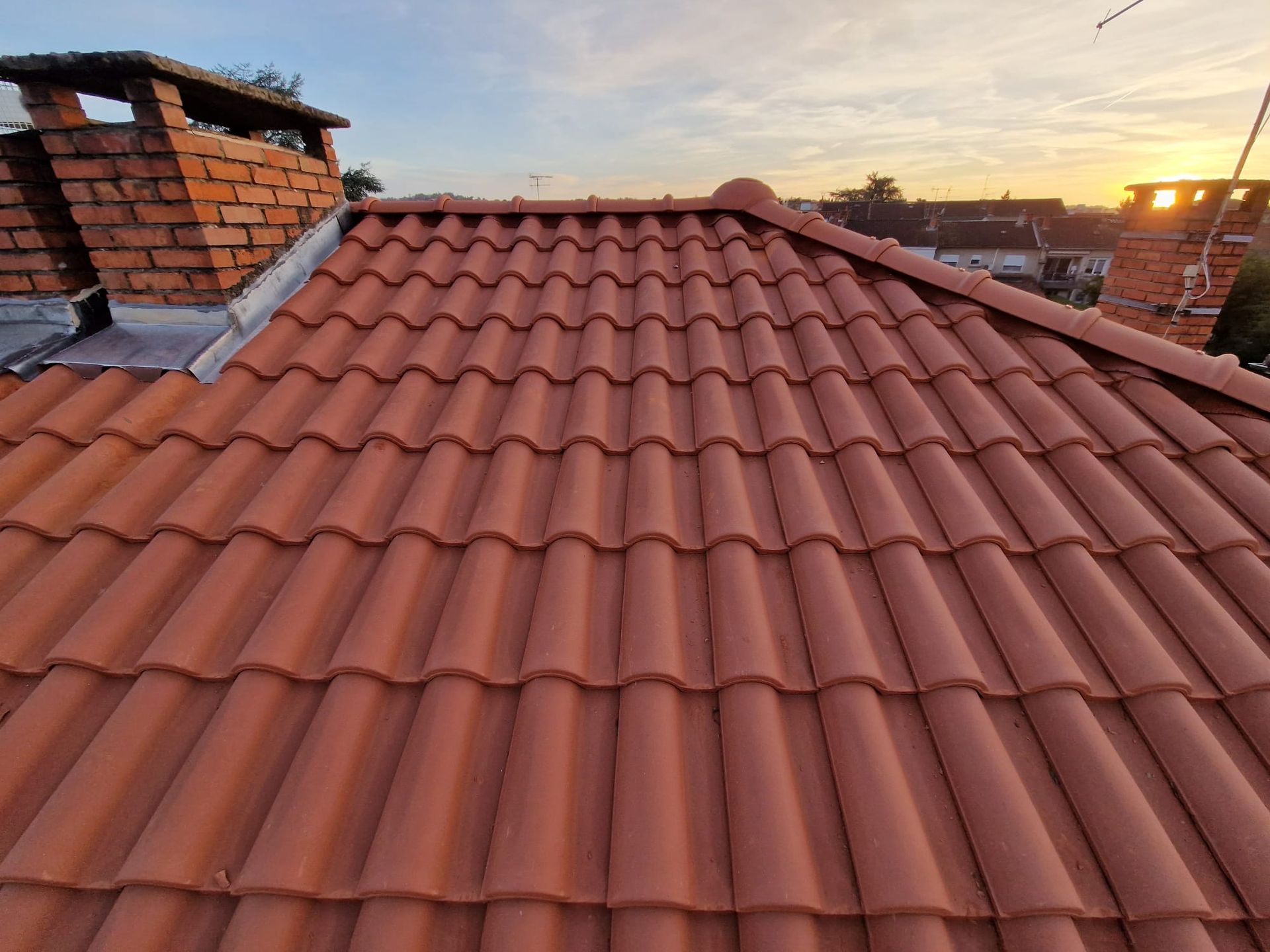 Toit de tuiles en terre cuite rouge avec deux cheminées en briques, sur fond de ciel de coucher de soleil.
