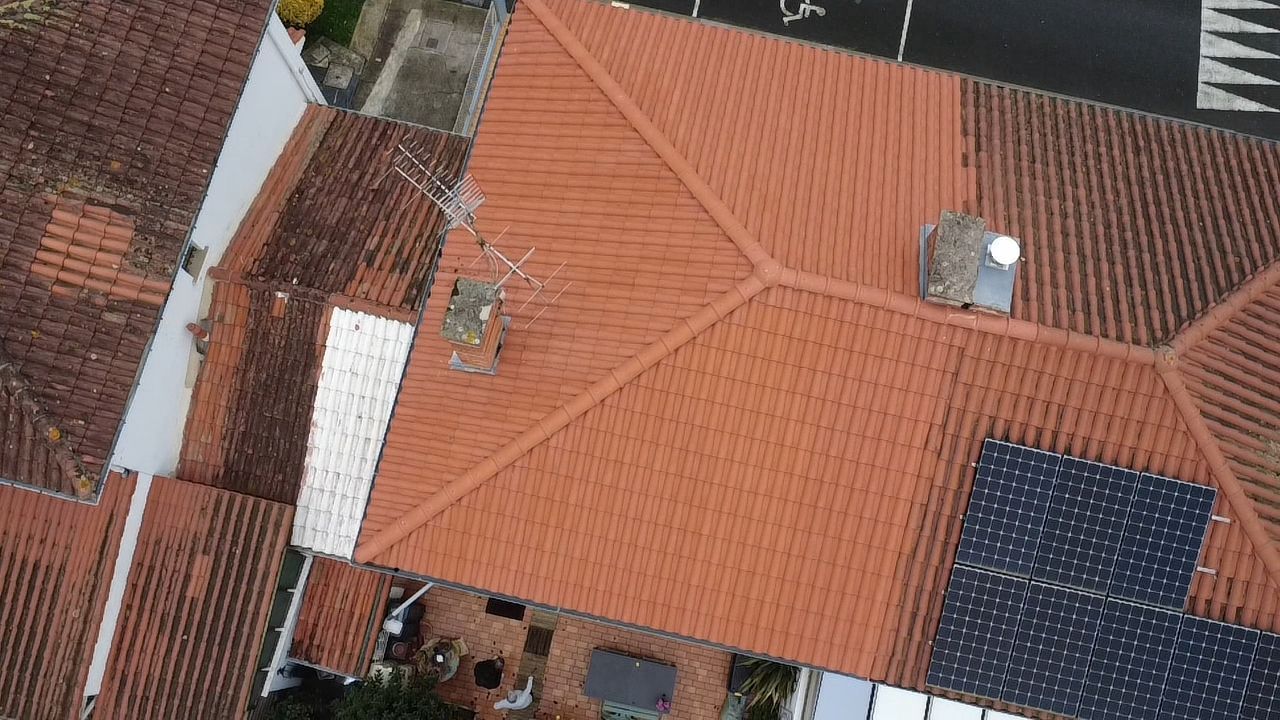 Vue aérienne d'un toit en tuiles de terre cuite comportant un ensemble de panneaux solaires, une cheminée et une antenne.