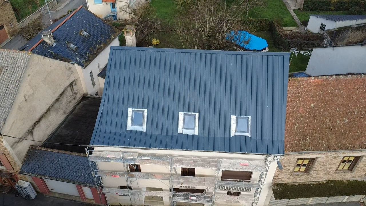 Vue aérienne d'un bâtiment en rénovation, présentant une nouvelle toiture métallique à joints debout bleu foncé avec trois puits de lumière.