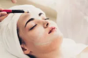 Se está aplicando una mascarilla facial a una persona recostada con un pincel y una diadema blanca.