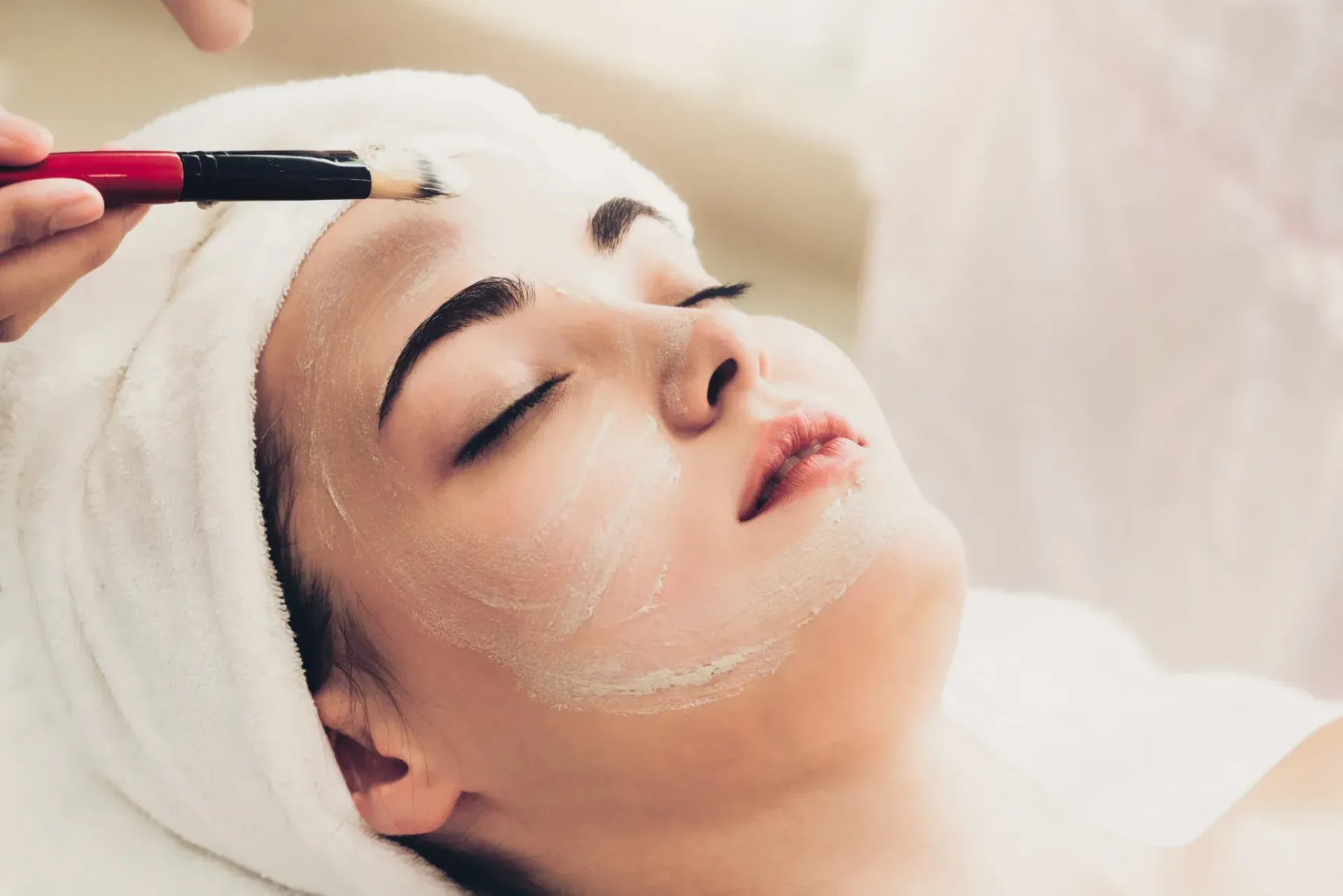 Se está aplicando una mascarilla facial a una persona recostada con un pincel y una diadema blanca.