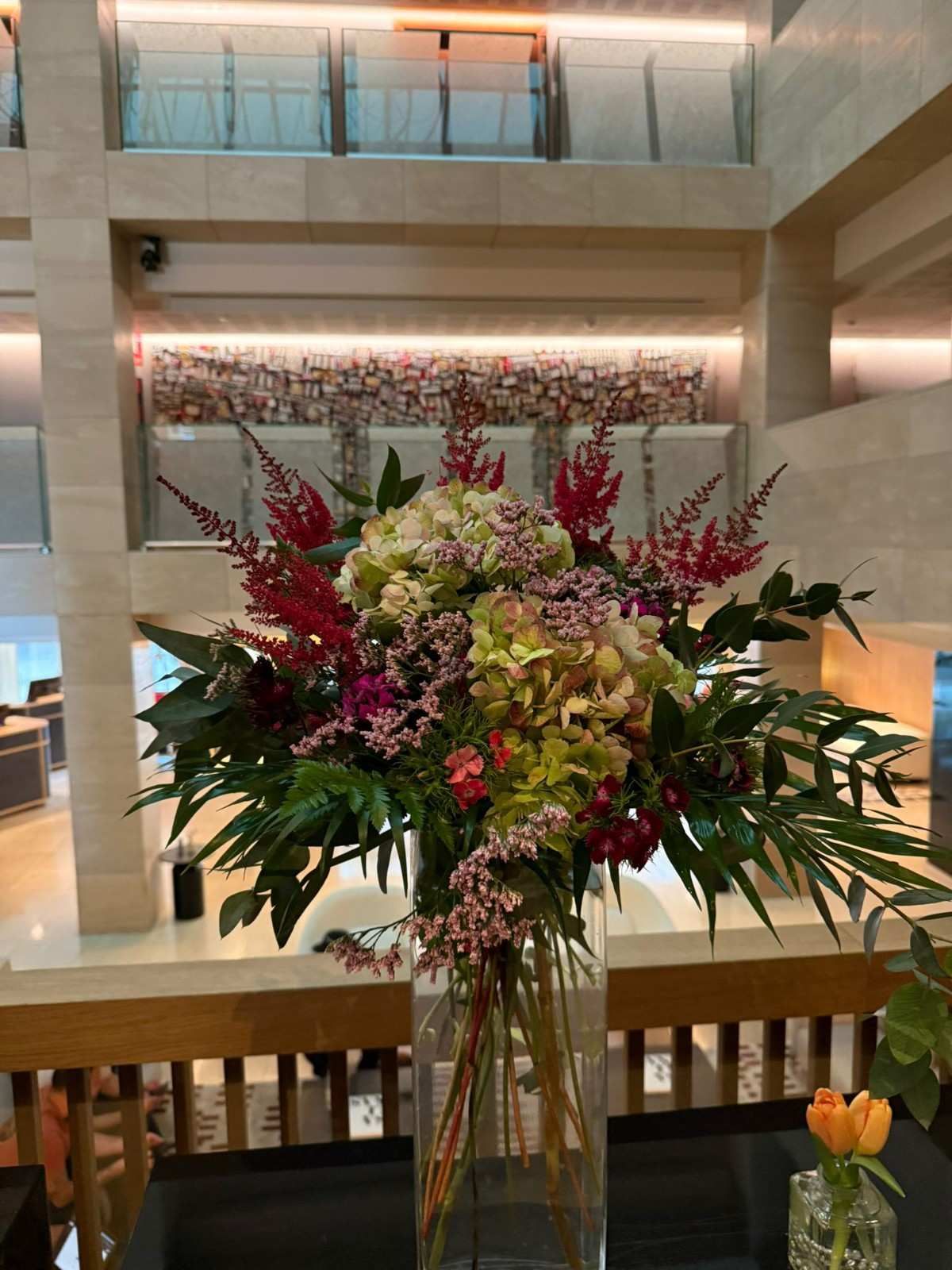 Un gran arreglo floral con hortensias verdes, astilbe rojo y follaje oscuro en un jarrón transparente en un atrio luminoso.