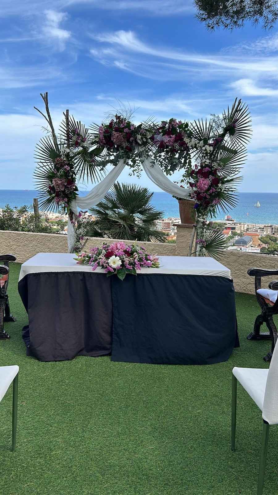 Un altar nupcial con arreglos florales y tela blanca drapeada, con vistas al mar en una soleada ladera cubierta de hierba.