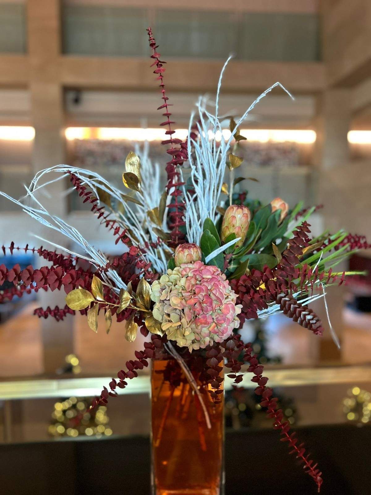 Un jarrón alto de cristal lleno de un ramo de hortensias rosas, hojas doradas, eucalipto rojo y ramas blancas de invierno.