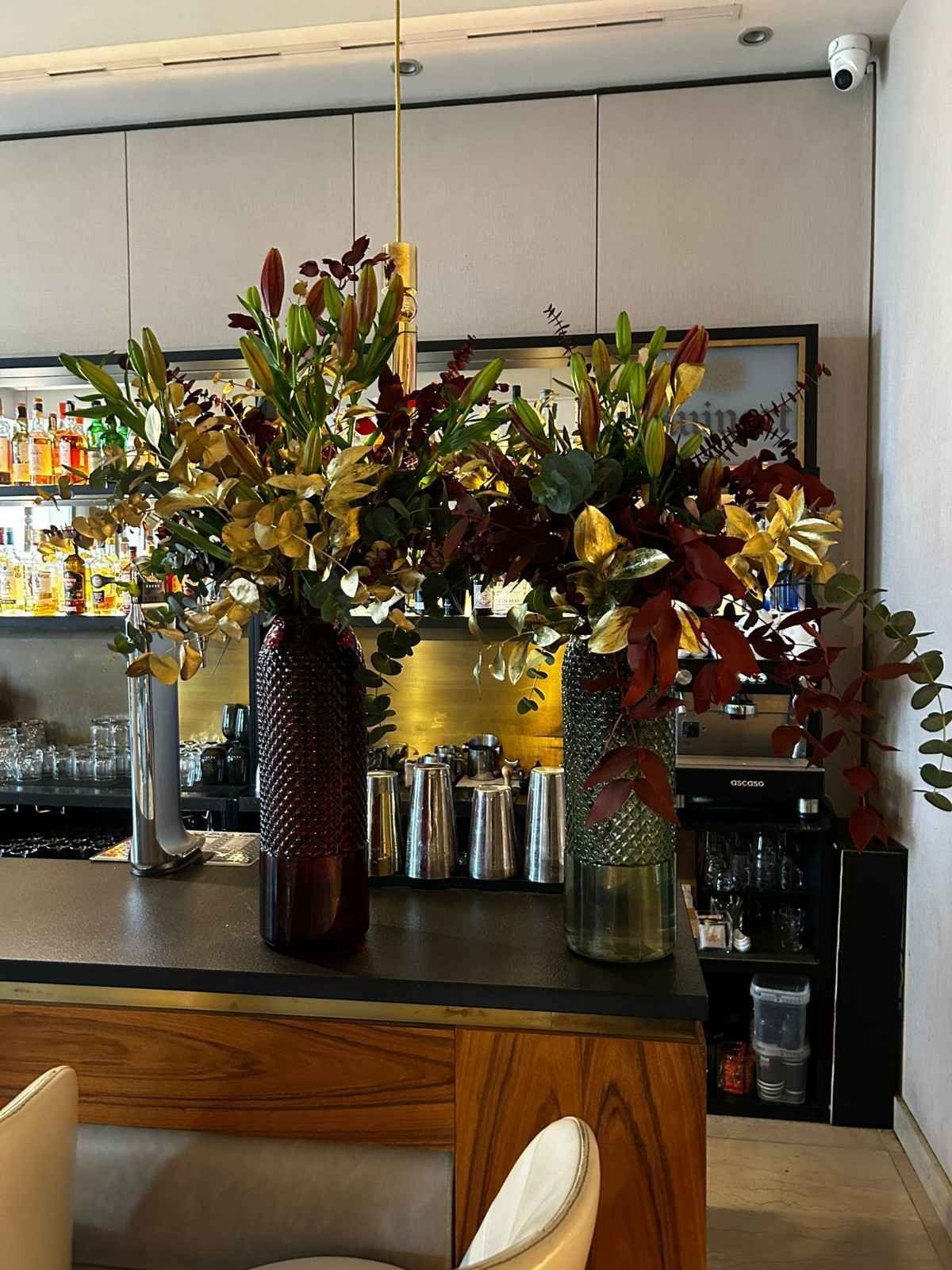 Dos jarrones altos y texturizados contienen exuberantes arreglos de lirios y follaje oscuro sobre la barra de un bar