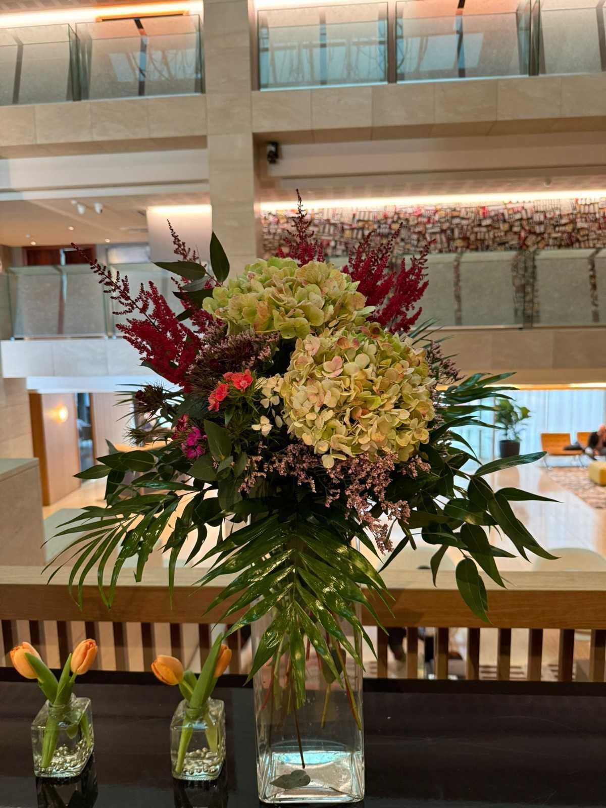 Un gran arreglo floral con hortensias y follaje reposa sobre una mesa cerca de dos jarrones
