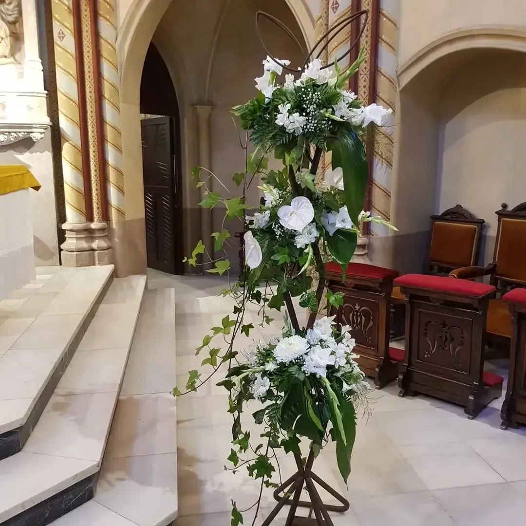 Un arreglo floral escalonado con flores blancas y hiedra sobre un soporte de metal oscuro en el interior de una iglesia.