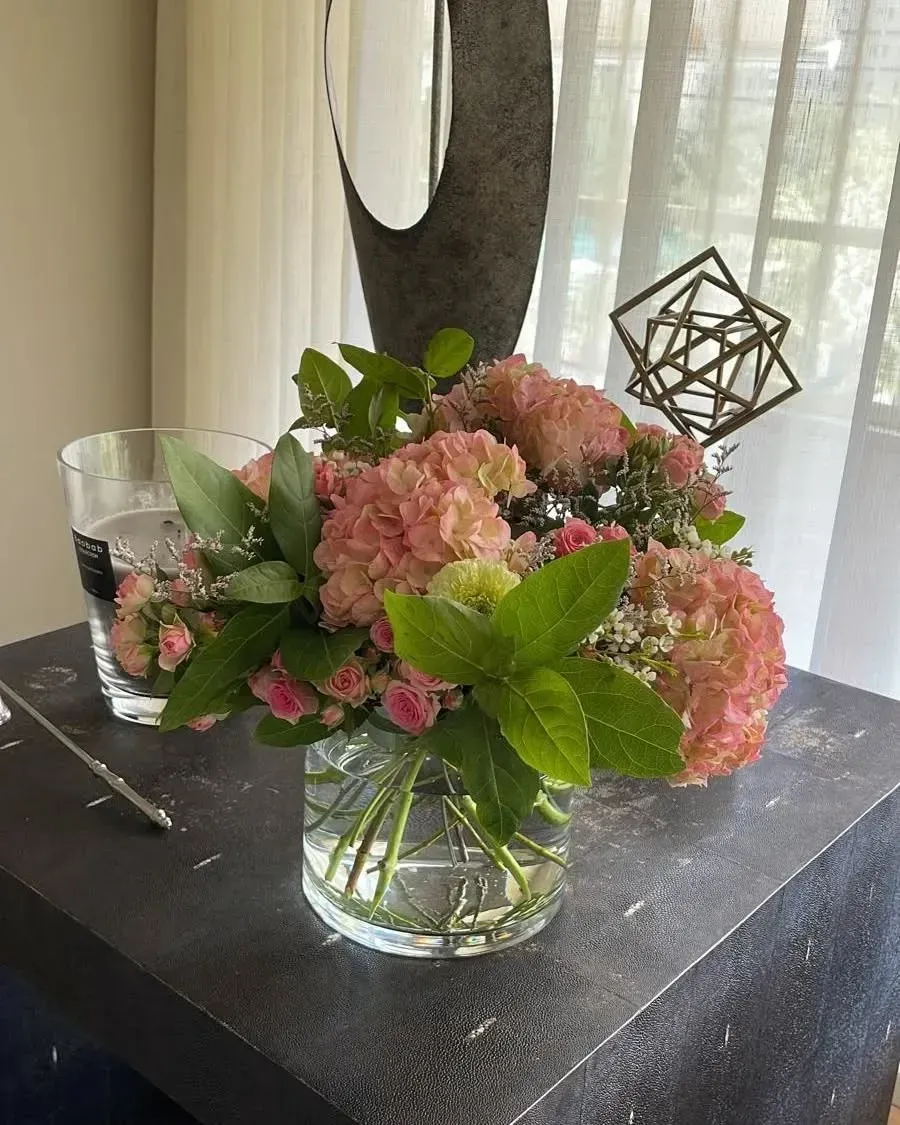Un jarrón de cristal lleno de hortensias rosas y pequeñas rosas reposa sobre una mesa oscura
