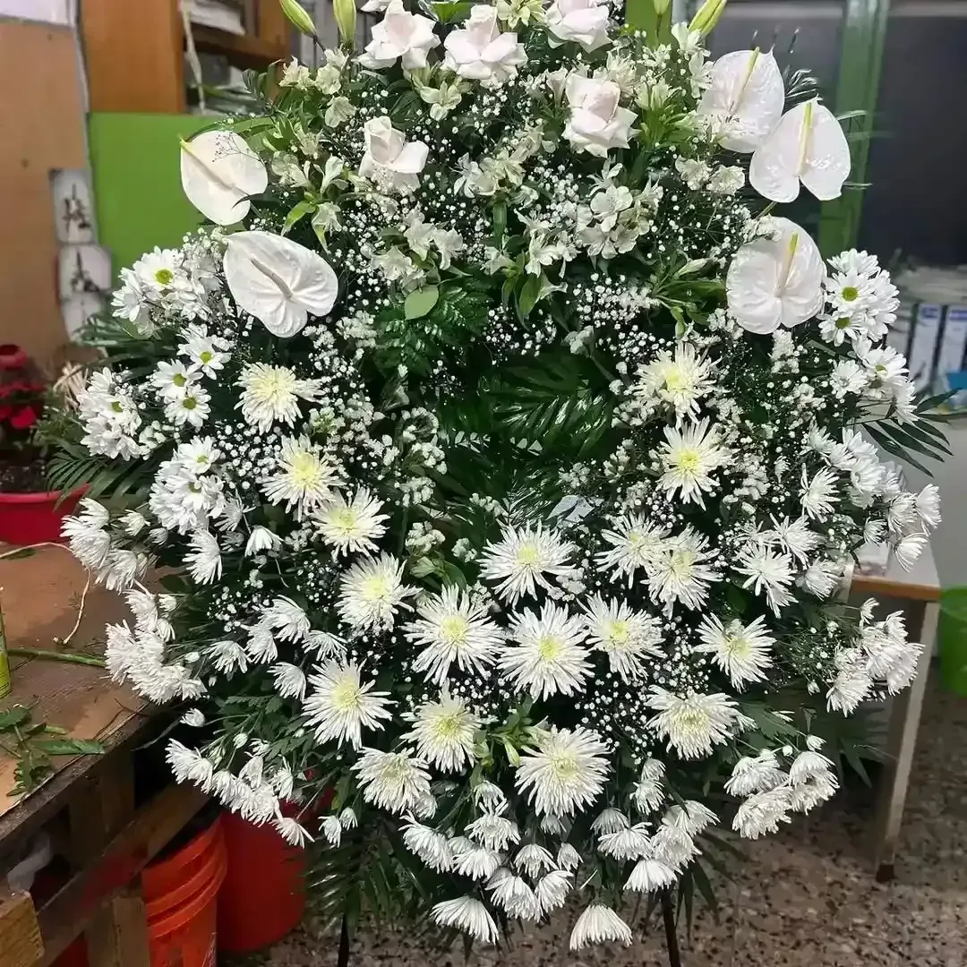Una corona funeraria compuesta de crisantemos blancos, anturios blancos y follaje, con un taller como fondo.