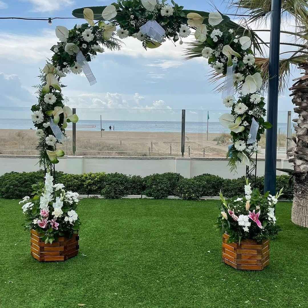Un arco decorado con flores se alza sobre un césped junto a la playa, adornado con flores blancas y vegetación