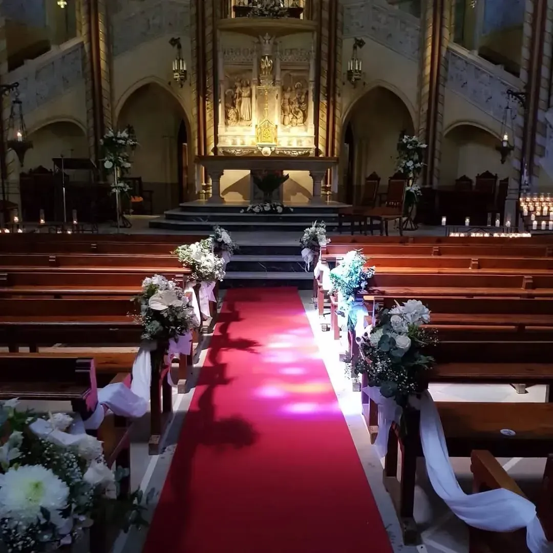Un pasillo alfombrado de rojo en el interior de una iglesia, adornado con ramos de flores sobre bancos de madera