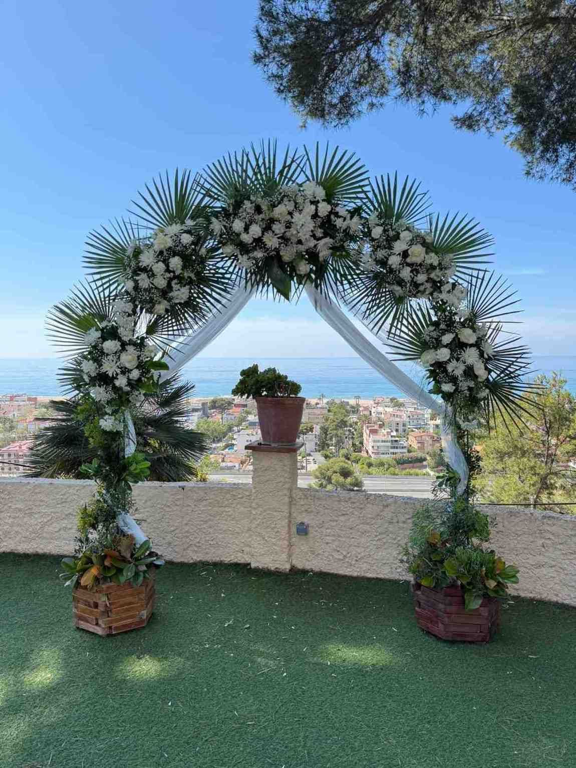 Un arco nupcial triangular blanco decorado con rosas blancas y hojas de palma, situado al aire libre