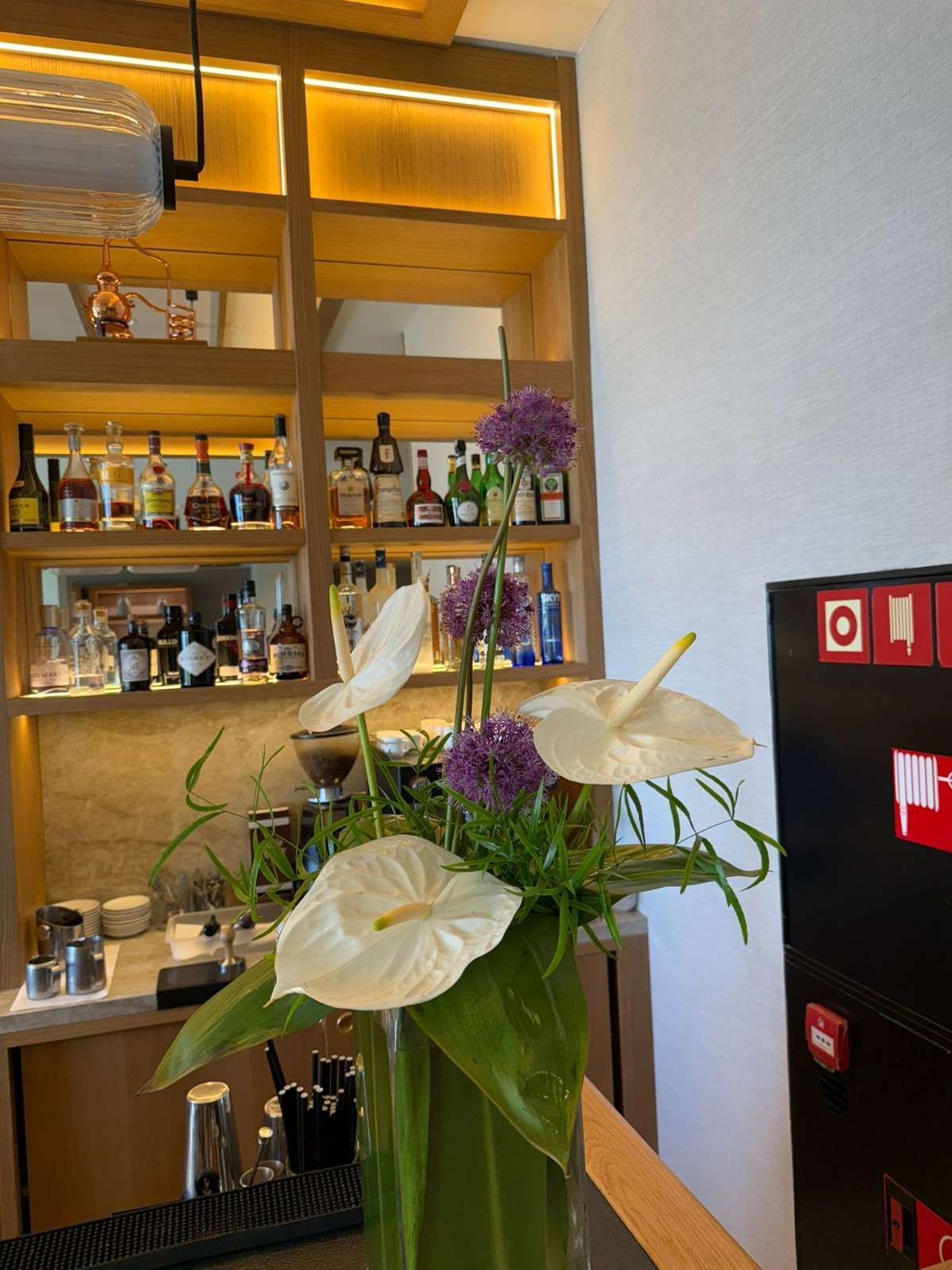 Un arreglo floral con anturios blancos y allium morados reposa sobre la barra de un bar con estantes llenos de botellas