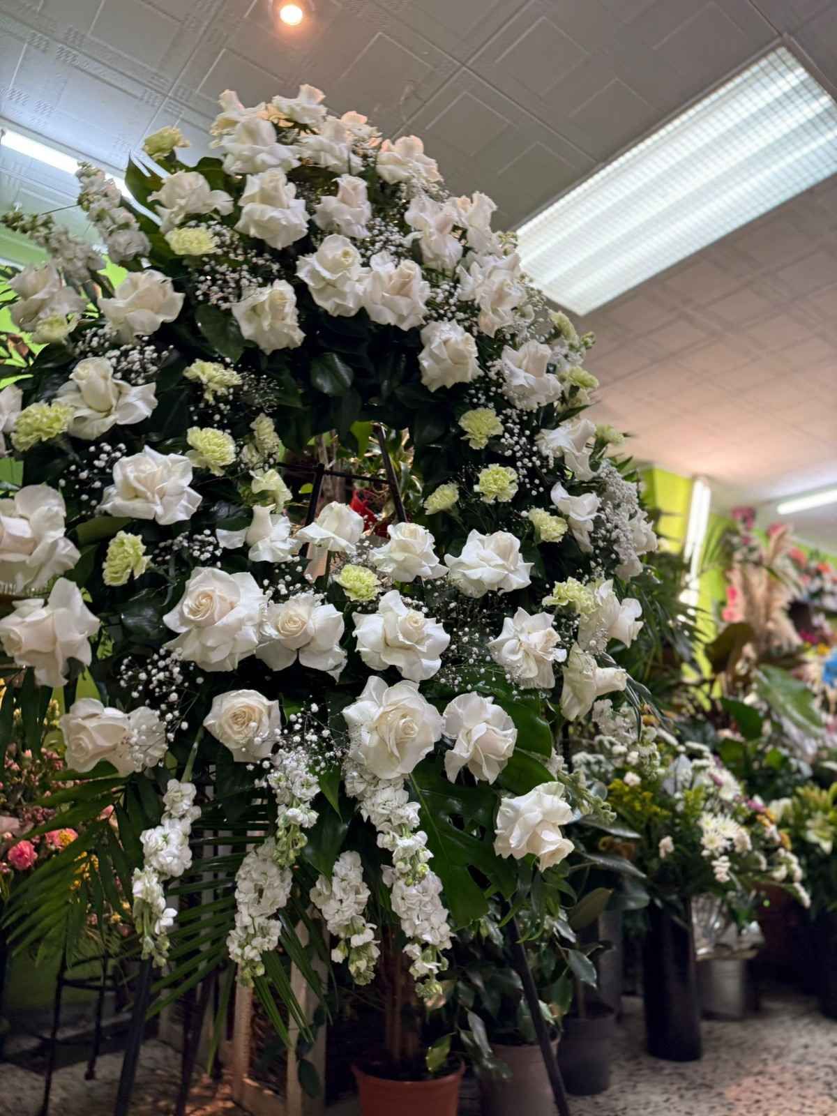 Una gran y exuberante corona funeraria hecha de rosas blancas y follaje verde sobre un pedestal dentro de una floristería.