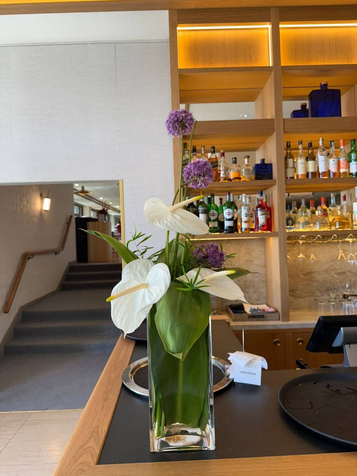 Un arreglo floral de anturios blancos y flores de allium moradas en un jarrón transparente sobre la barra de un bar