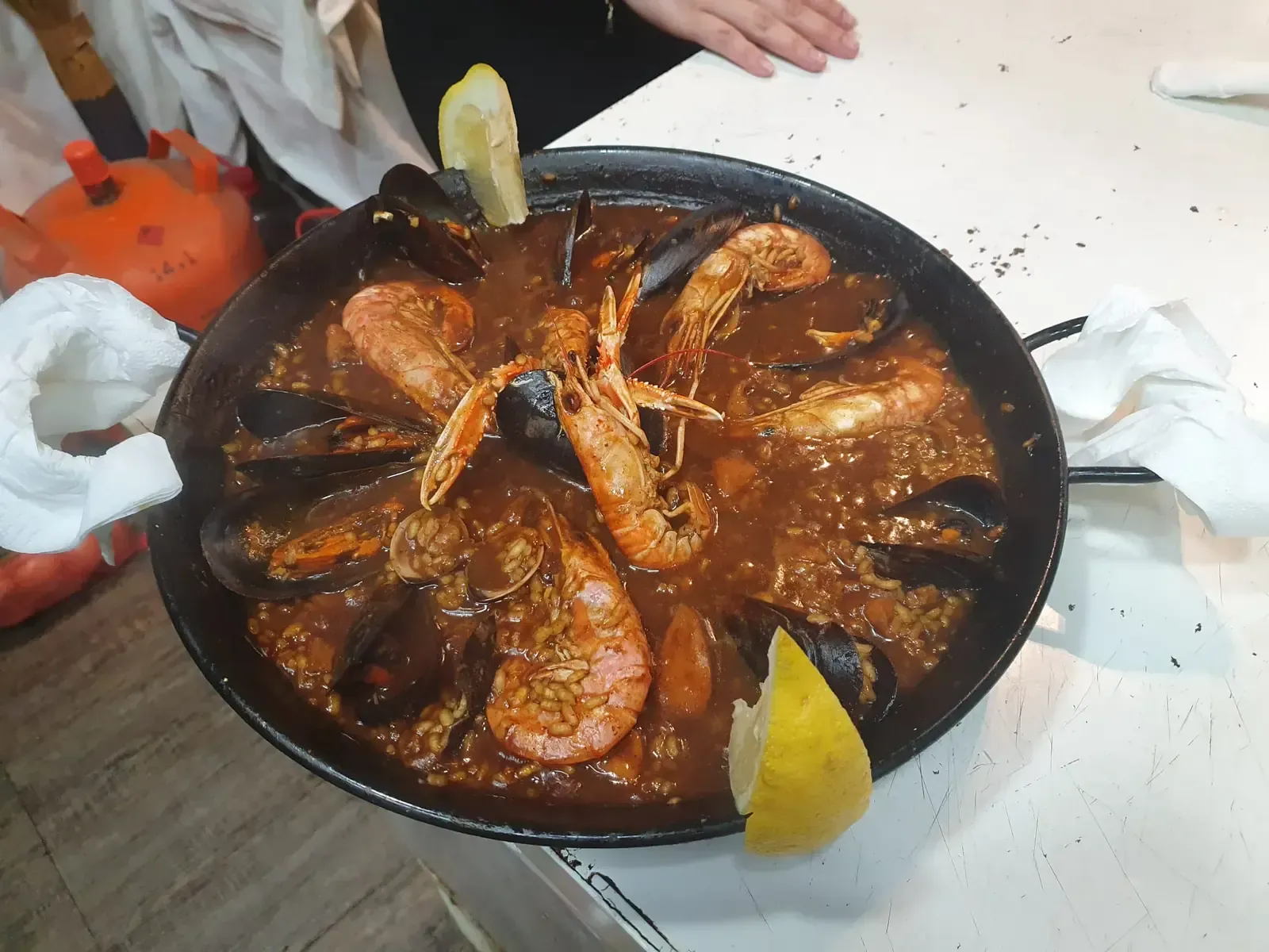 Sartén con gambas cocidas en salsa oscura y rodajas de limón sobre una mesa; se ven manos cerca.