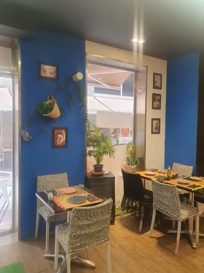 Interior del restaurante con paredes azules, mesas pequeñas, sillas de mimbre y una ventana con vistas al exterior.