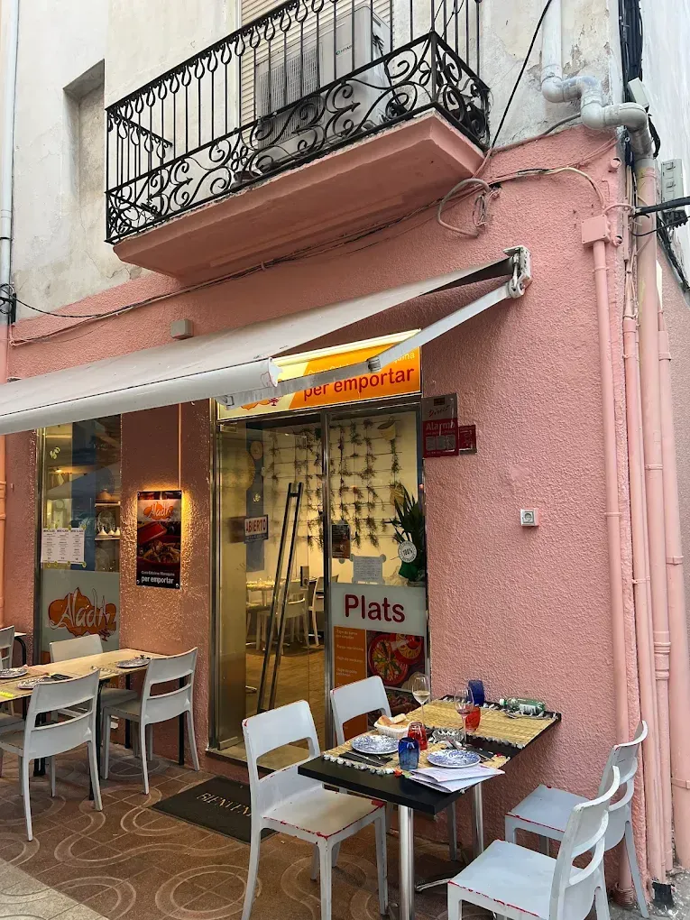 Cafetería al aire libre con fachada rosa, mesas pequeñas, sillas y un balcón sobre la entrada.