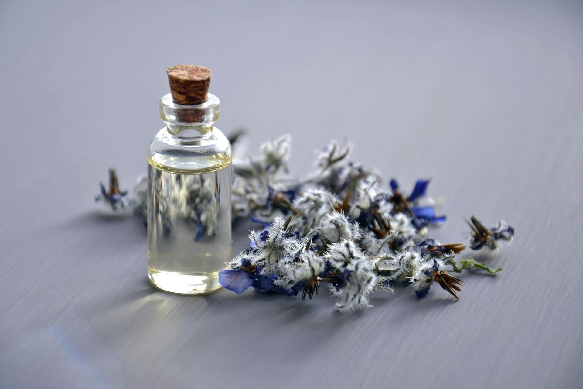 Pequeña botella de vidrio con aceite junto a flores secas azules y blancas sobre una superficie gris.