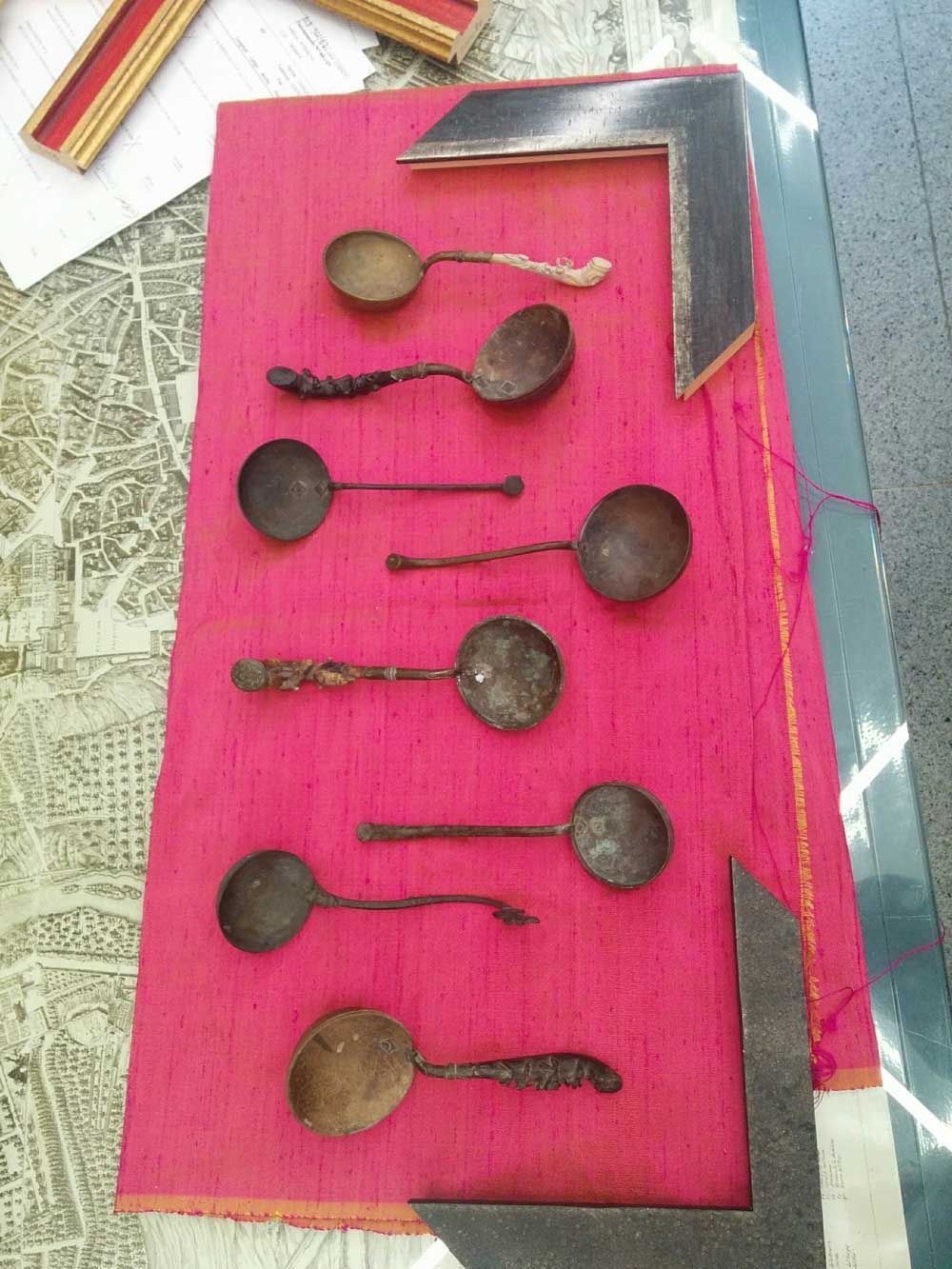 Exposición de cucharas antiguas sobre fondo rosa, enmarcadas por herramientas.