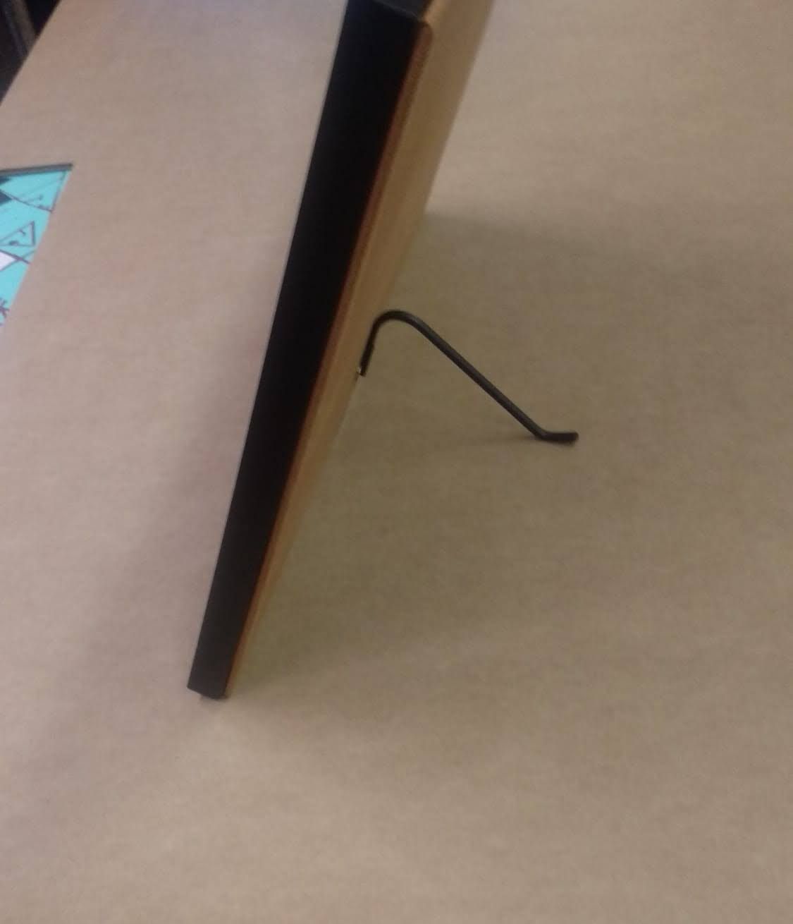 Cuadro enmarcado en negro, apoyado sobre un soporte de alambre, sobre una superficie de color tostado.