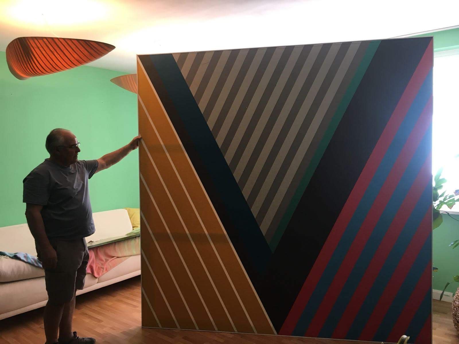 Un hombre se encuentra junto a una gran pintura abstracta con rayas diagonales de colores en un entorno doméstico.