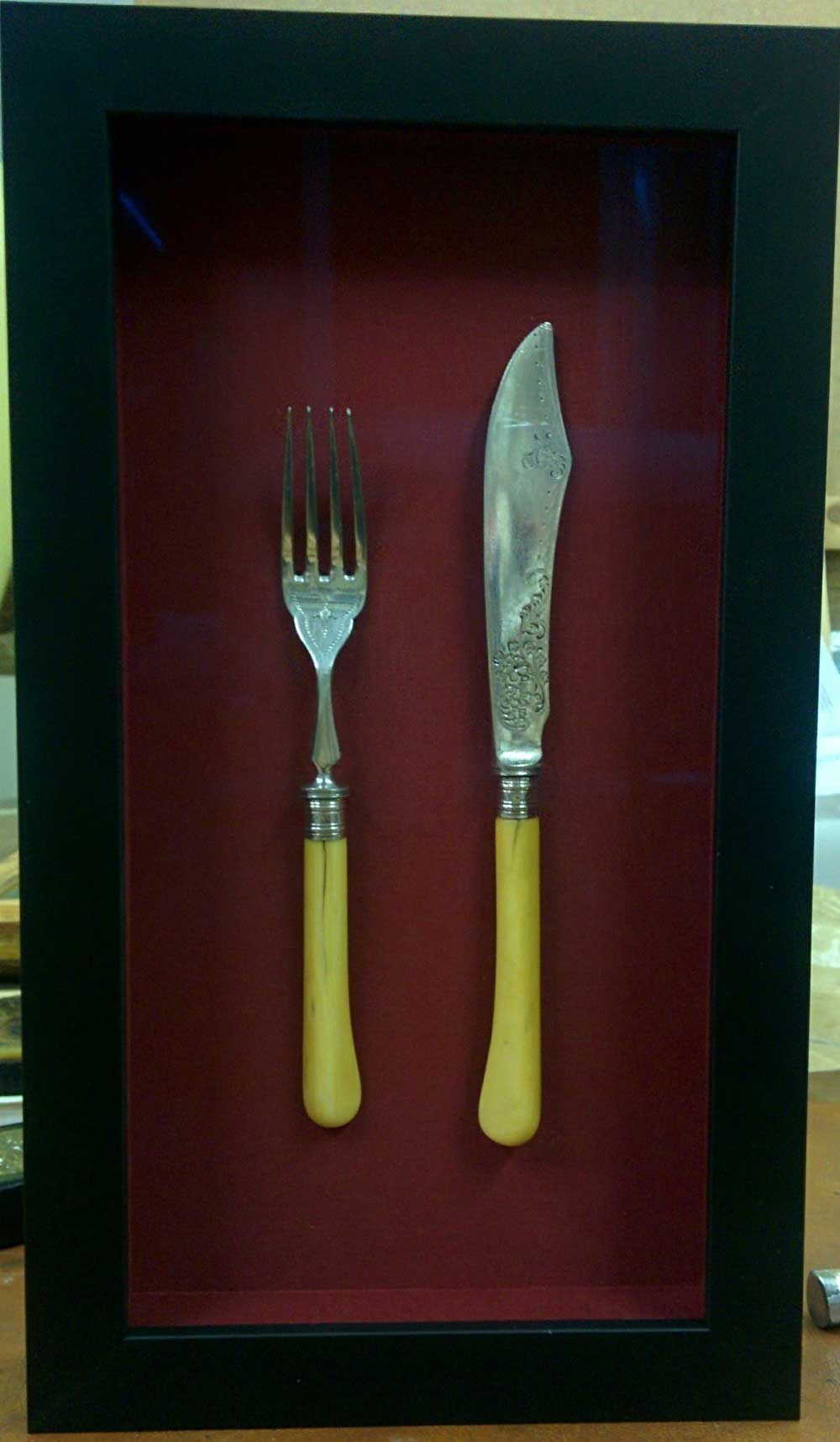 Juego de tenedor y cuchillo con mangos claros en una caja de sombra sobre un fondo rojo.