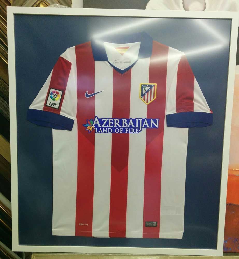 Camiseta del Atlético de Madrid enmarcada; rayas rojas y blancas, patrocinador Azerbaiyán, marco blanco sobre fondo azul.