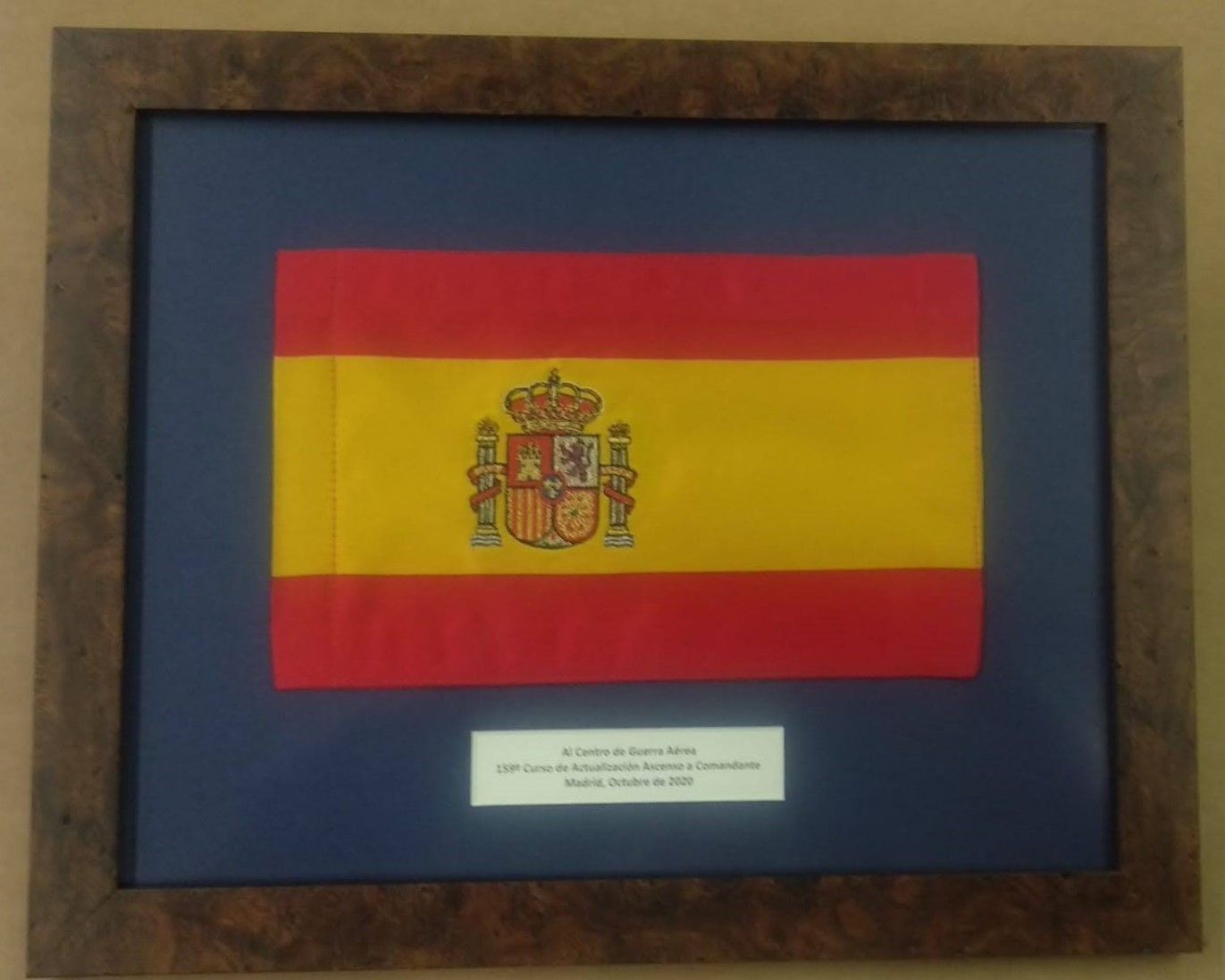 Bandera de España enmarcada en madera oscura, expuesta sobre fondo azul, con una pequeña leyenda.