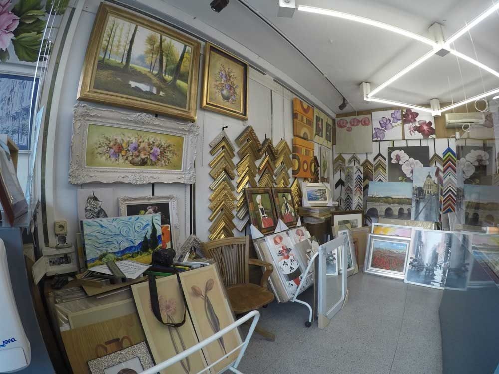 Interior de galería de arte, pinturas y marcos expuestos en paredes y mesas, iluminación brillante.