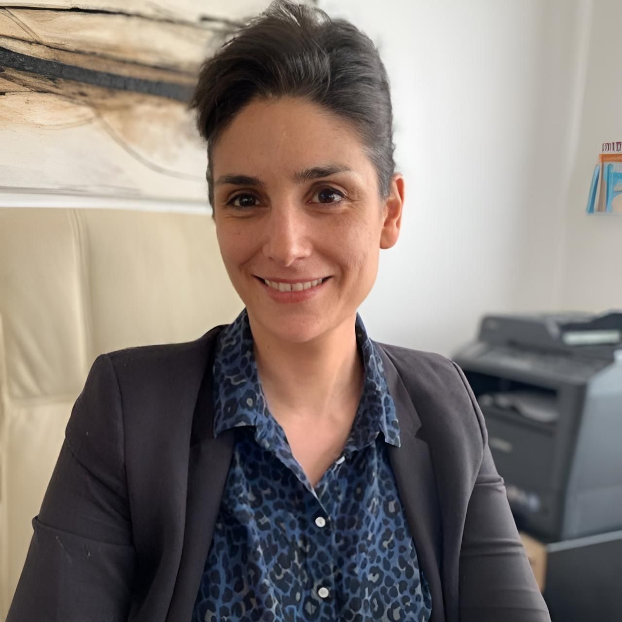 Une femme en blazer et chemise à motifs sourit, assise dans un bureau, avec une imprimante visible en arrière-plan.